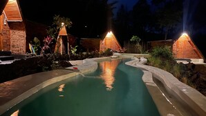 Ein Resort bei Nacht mit Schwimmbad, Holzhütten und tropischer Bepflanzung.