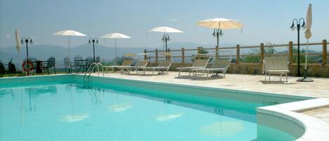 Piscina con sillas de descanso y sombrillas, rodeada por un suelo de piedra y una valla de madera.