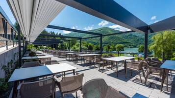 An outdoor patio with tables and chairs, overlooking a lake and mountains.