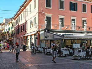 Eine Kopfsteinpflasterstraße gesäumt von Gebäuden, mit Außenplätzen eines Cafés und Menschen, die zu Fuß gehen und Fahrrad fahren.