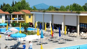 Un espace piscine avec des chaises longues et des parasols, un bâtiment doté de balcons, et une montagne en arrière-plan.