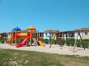 Ein Spielplatz mit Rutschen und Klettergerüsten, umgeben von Häusern mit Balkonen und Sonnenschirmen.