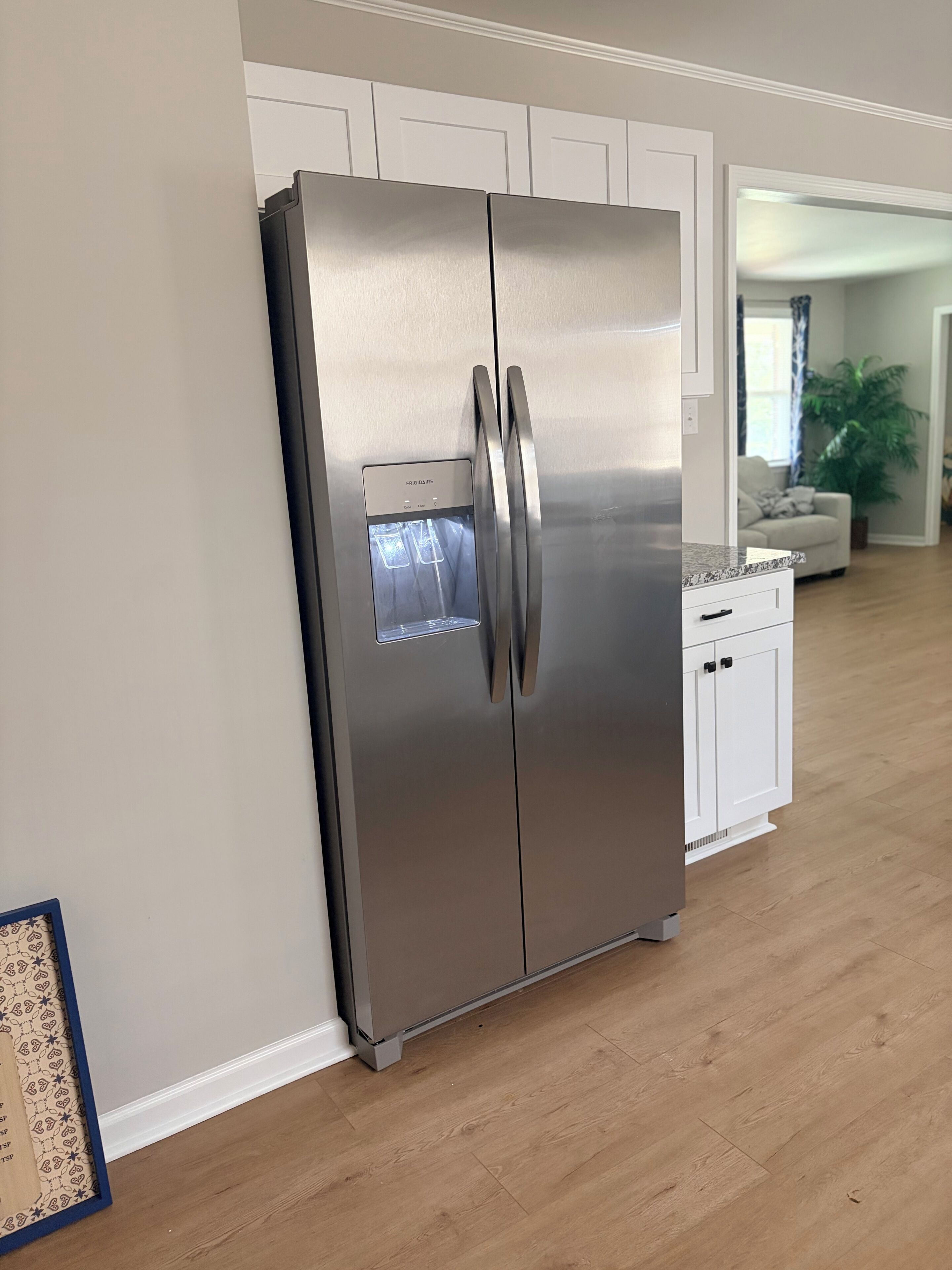 A stainless steel refrigerator with a water dispenser in a kitchen with white cabinetry and a wooden floor.