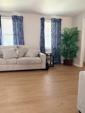 Una sala de estar con un sofá color beige, una mesita con una planta y un ventanal con cortinas.