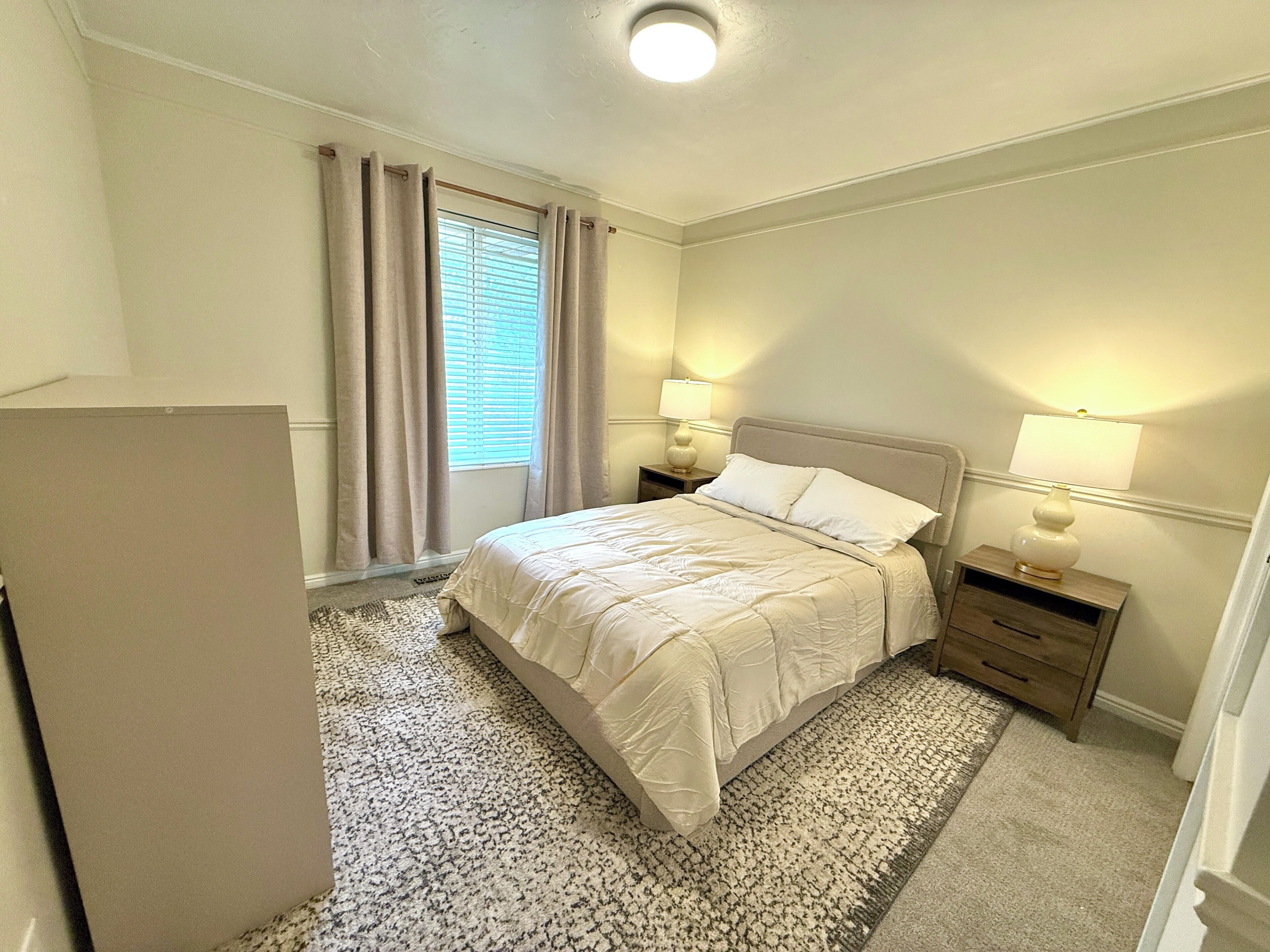 A bedroom with a bed, two bedside tables with lamps, a window with curtains, and a carpeted floor.