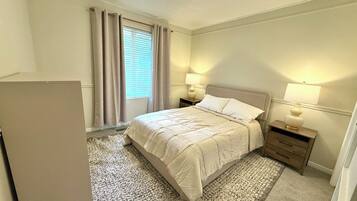 A bedroom with a bed, two bedside tables with lamps, a window with curtains, and a carpeted floor.