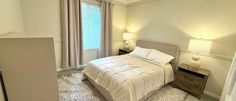 A bedroom with a bed, two bedside tables with lamps, a window with curtains, and a carpeted floor.