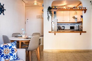 A dining area with a round table, grey chairs, and a decorative pillow. A kitchen with a wooden countertop and a hanging plant is visible through an archway. - Pink Buganvilla Lodging (Torrevieja)