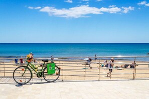 Une plage avec un vélo garé sur la promenade, des gens qui prennent un bain de soleil et l’océan en arrière-plan.