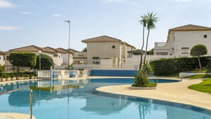 Uma piscina com superfície de água azul cristalina, cercada por uma cerca branca, e adjacente a uma fileira de edifícios com arquitetura semelhante.