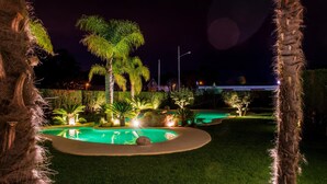 A nighttime view of a landscaped area with a pool, palm trees, and well-lit pathways. - Hotel Dinajan (Vilanova de Arousa)
