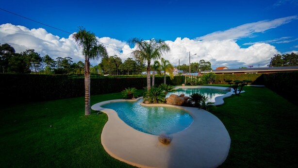 A swimming pool with a curved design, surrounded by well-maintained grass and palm trees. - Hotel Dinajan (Vilanova de Arousa)