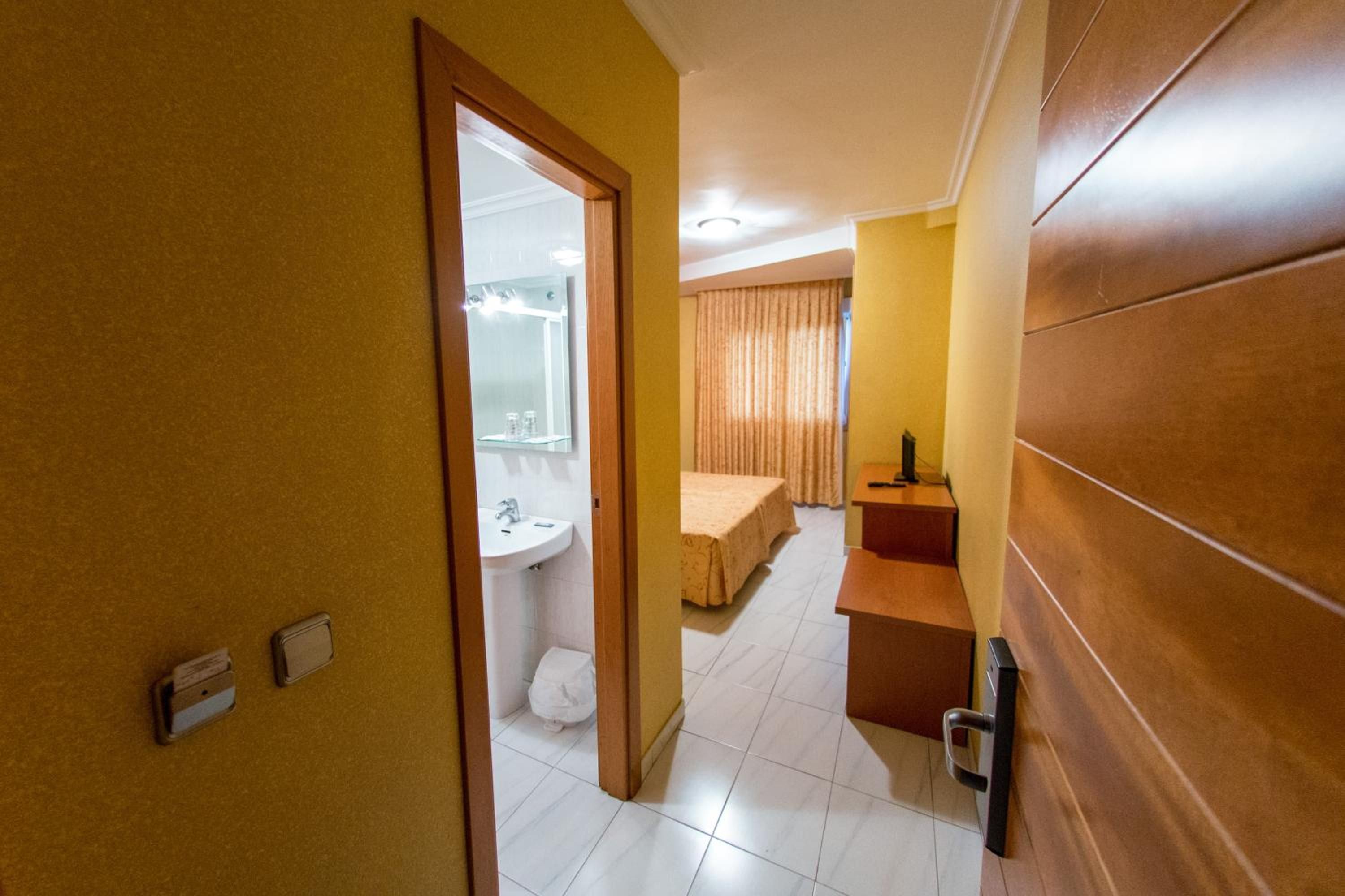 Una habitación de hotel con puerta de madera, baño con lavamanos y espejo, cama con cabecera y una mesita con lámpara.