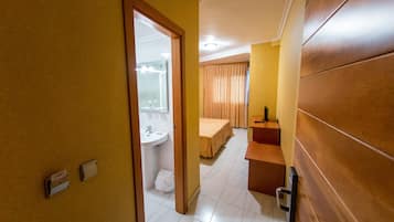Una habitación de hotel con puerta de madera, baño con lavamanos y espejo, cama con cabecera y una mesita con lámpara.
