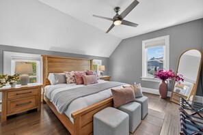 A bedroom with a bed, bedside tables, a ceiling fan, a window with a view, a mirror, and a vase with flowers. - Lavender Hill Skyline - Sleeps 12 - City Views (Milwaukee)