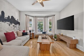 A modern living room with a sofa, coffee table, and a flat-screen TV mounted on the wall. - Lavender Hill Skyline - Sleeps 12 - City Views (Milwaukee)