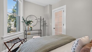 Ein Schlafzimmer mit Bett, Fenster, Kommode, Spiegel und Tür.