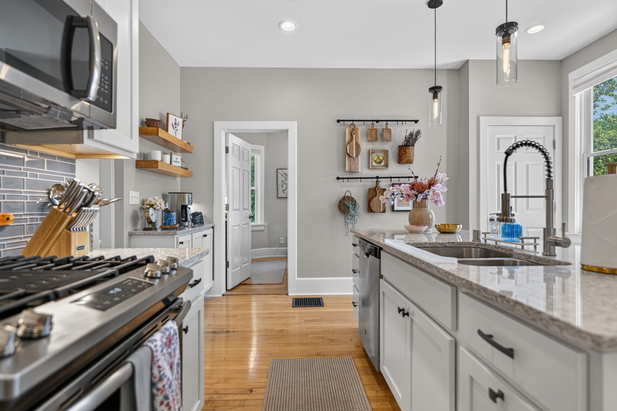 A modern kitchen with a stainless steel stove, a microwave, and a sink with a faucet. There are wooden shelves above the stove and a hanging rack with kitchen utensils.