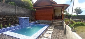 Una piscina con borde de azulejos, una estructura de madera con porche y una pared de piedra.