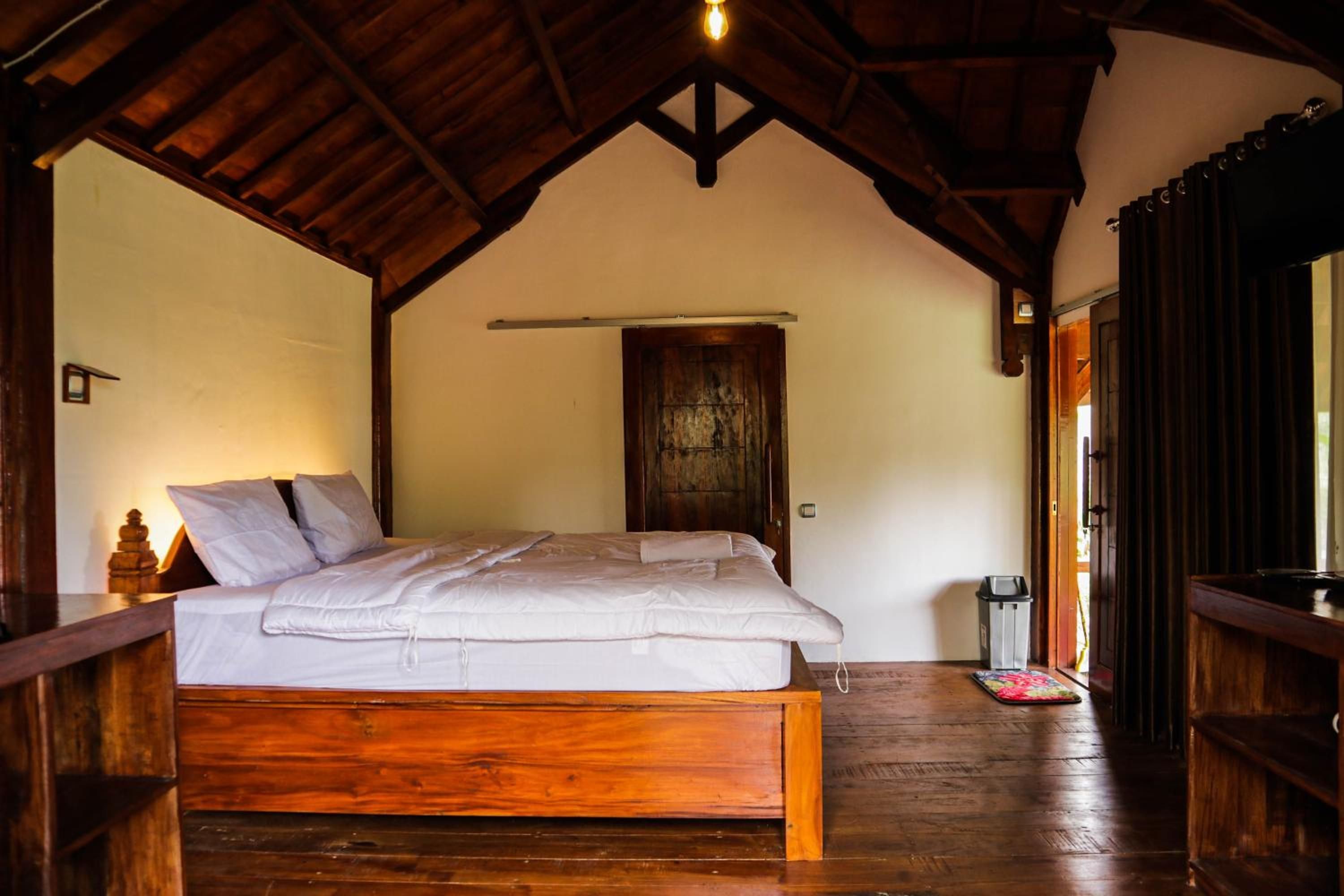 A wooden-framed bedroom with a bed, a wooden door, and a small trash bin.