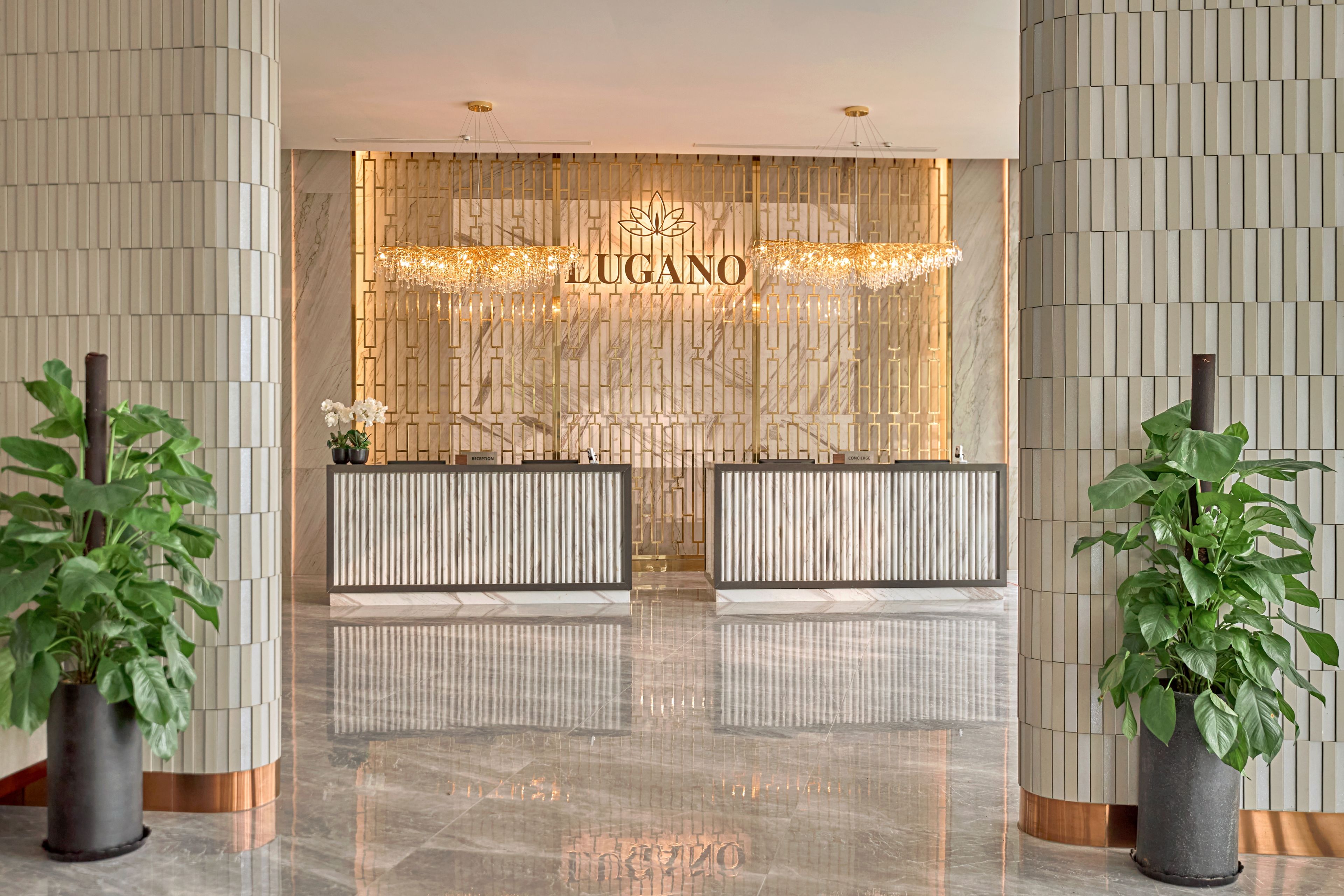 Zona de recepción de un hotel con piso de mármol, dos mostradores identificados como 'RECEPTION' y 'CONCIERGE', y una pared decorativa con el nombre del hotel 'LUGANO'.