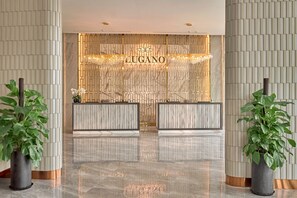 Ein Hotel-Rezeptionsbereich mit Marmorboden, zwei Empfangstresen, beschriftet mit 'RECEPTION' und 'CONCIERGE', sowie einer Wand mit dem Hotelnamen 'LUGANO'.