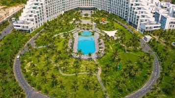 Un hotel grande con una piscina central rodeada de palmeras y una carretera curva.
