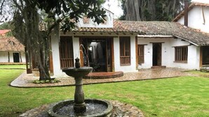A courtyard with a stone fountain, a white building with a tiled roof, and a tree. - Hacienda La Armenia - Cottage Hotel (Sopó)