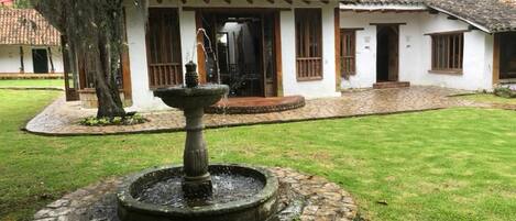 Un patio con una fuente de piedra, un edificio blanco con techo de tejas y un árbol.