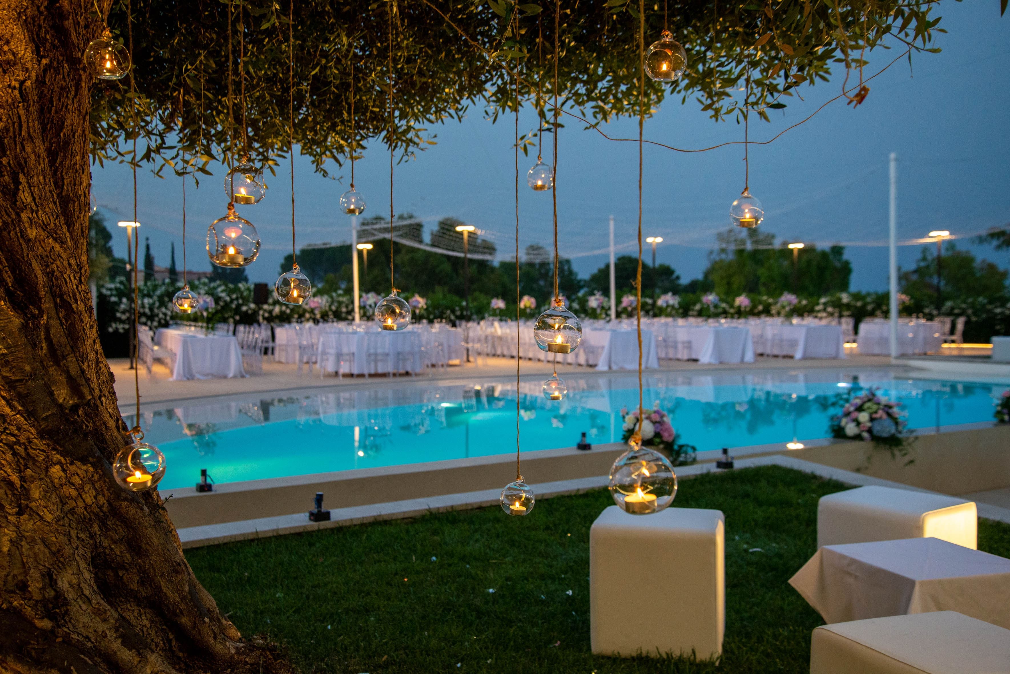An outdoor pool area with hanging glass orbs containing candles, surrounded by tables with white tablecloths.