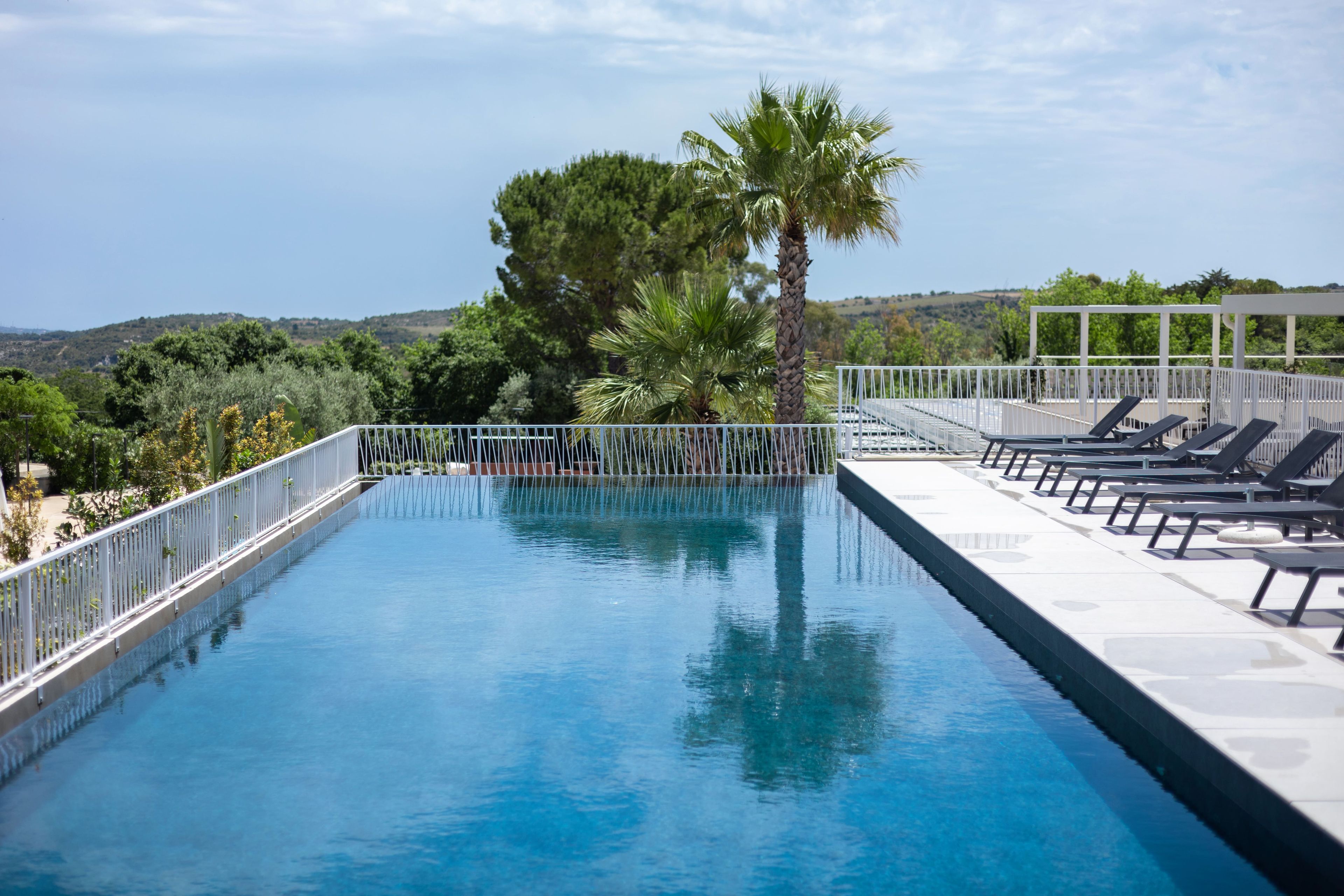 A rectangular swimming pool with a white railing, surrounded by lounge chairs and palm trees, with a view of hills in the background.