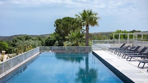 A rectangular swimming pool with a white railing, surrounded by lounge chairs and palm trees, with a view of hills in the background. - Tenuta San Marco (Noto)