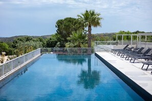 A rectangular swimming pool with a white railing, surrounded by lounge chairs and palm trees, with a view of hills in the background. - Tenuta San Marco (Noto)