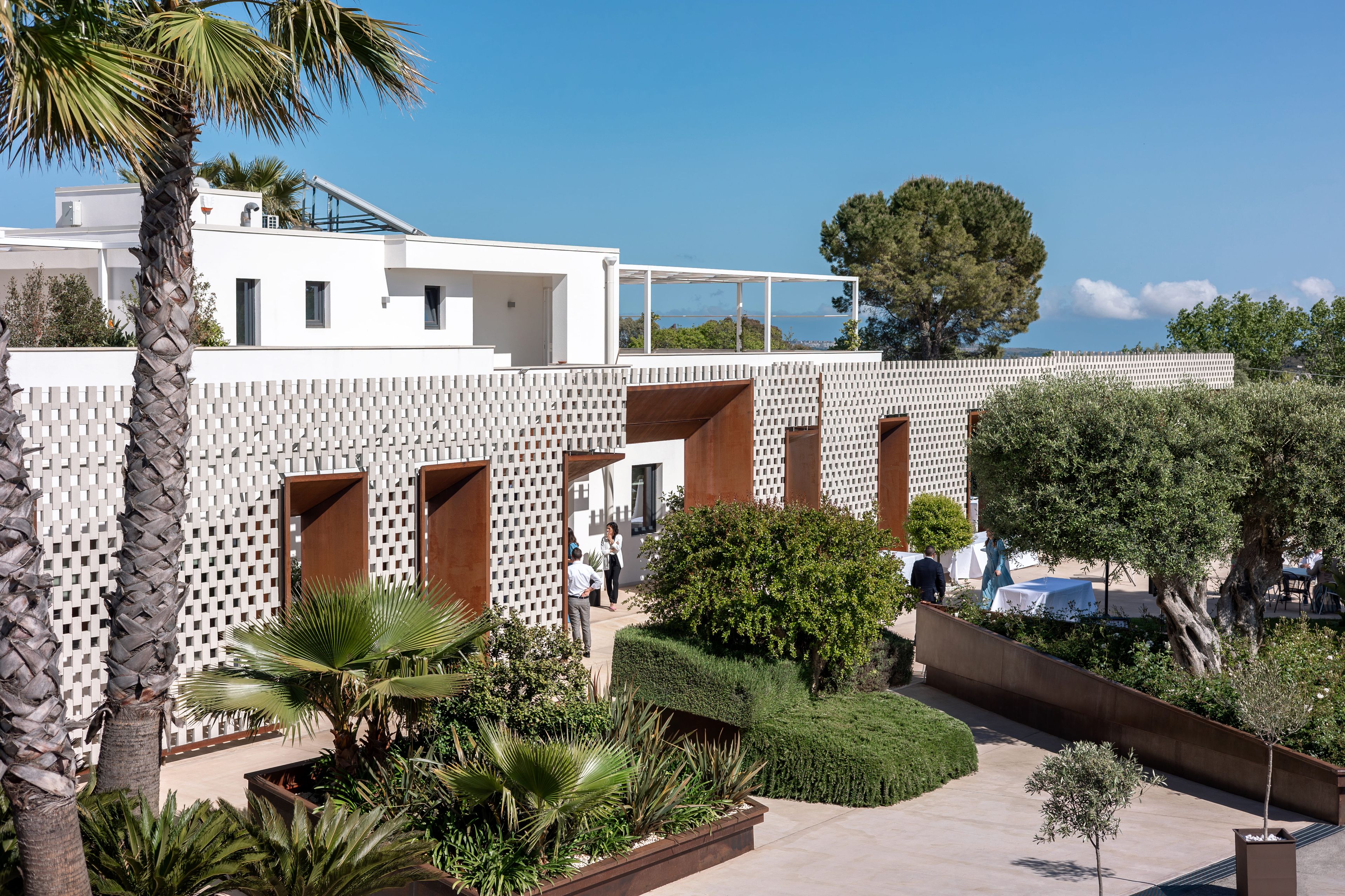 A modern building with a white facade, multiple balconies, and a landscaped garden with palm trees and shrubs.