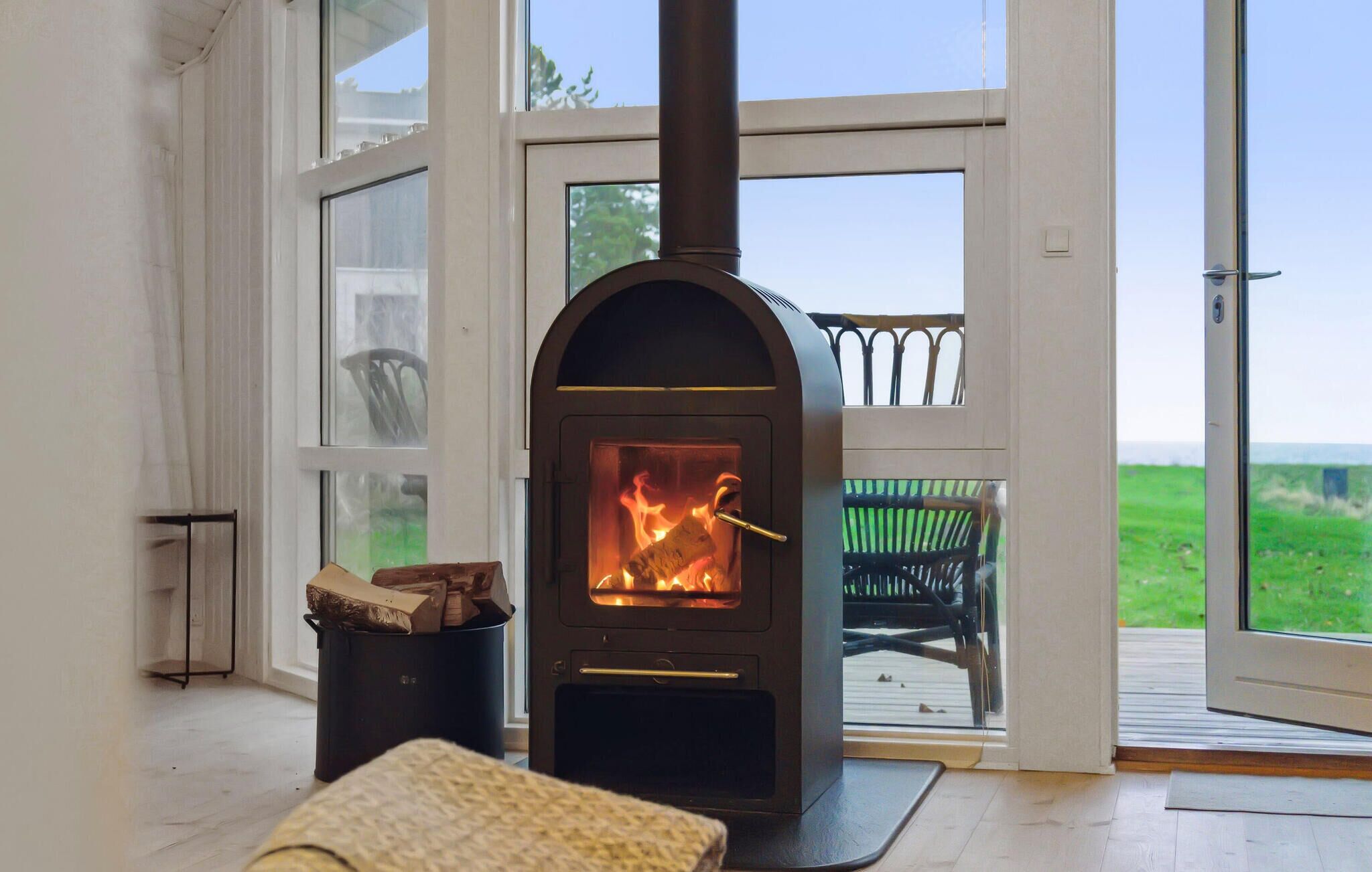 En vedeldad kamin med en glaslucka, en svart soptunna och en flätstol på ett trädäck.