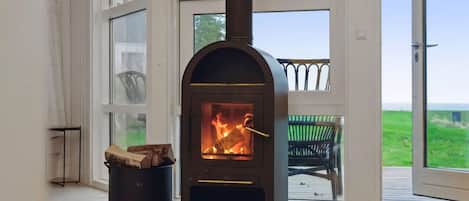 En vedeldad kamin med en glaslucka, en svart soptunna och en flätstol på ett trädäck.