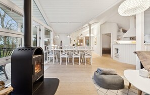 A modern interior with a wood-burning stove, a dining area with white chairs, and a kitchen with white cabinetry. - 3 bedroom stunning home in Martofte (Martofte)