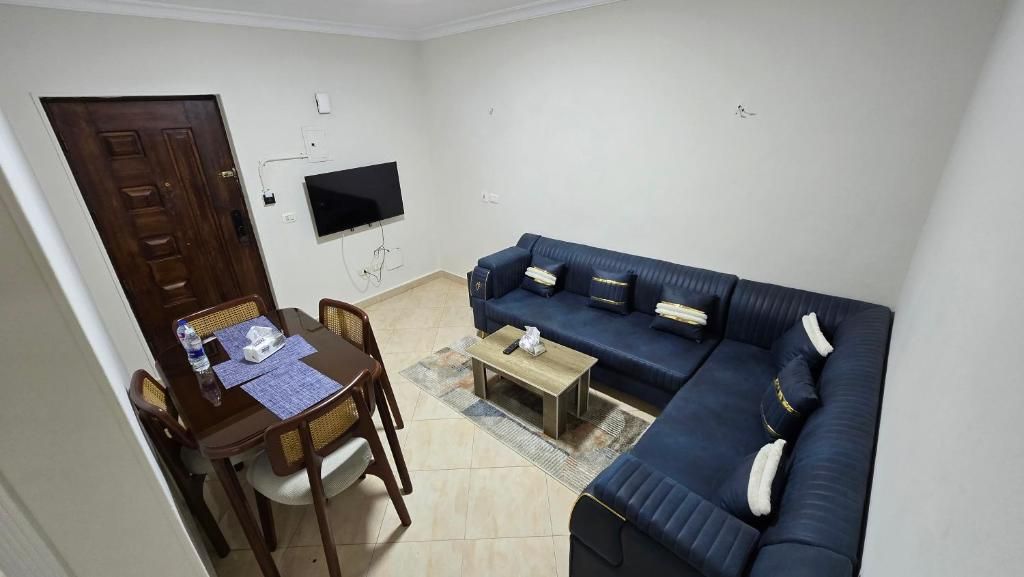 A living room with a dark blue sofa, a wooden dining table with chairs, a small coffee table, and a flat-screen TV mounted on the wall.