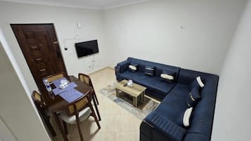A living room with a dark blue sofa, a wooden dining table with chairs, a small coffee table, and a flat-screen TV mounted on the wall.