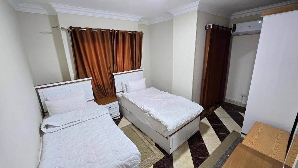 Habitación de hotel con dos camas individuales, una mesita de noche de madera, un armario blanco y suelo a cuadros.
