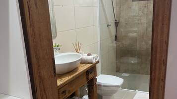 Casa de banho com lavatório branco, toucador de madeira, sanita branca e cabine de duche com parede de vidro.