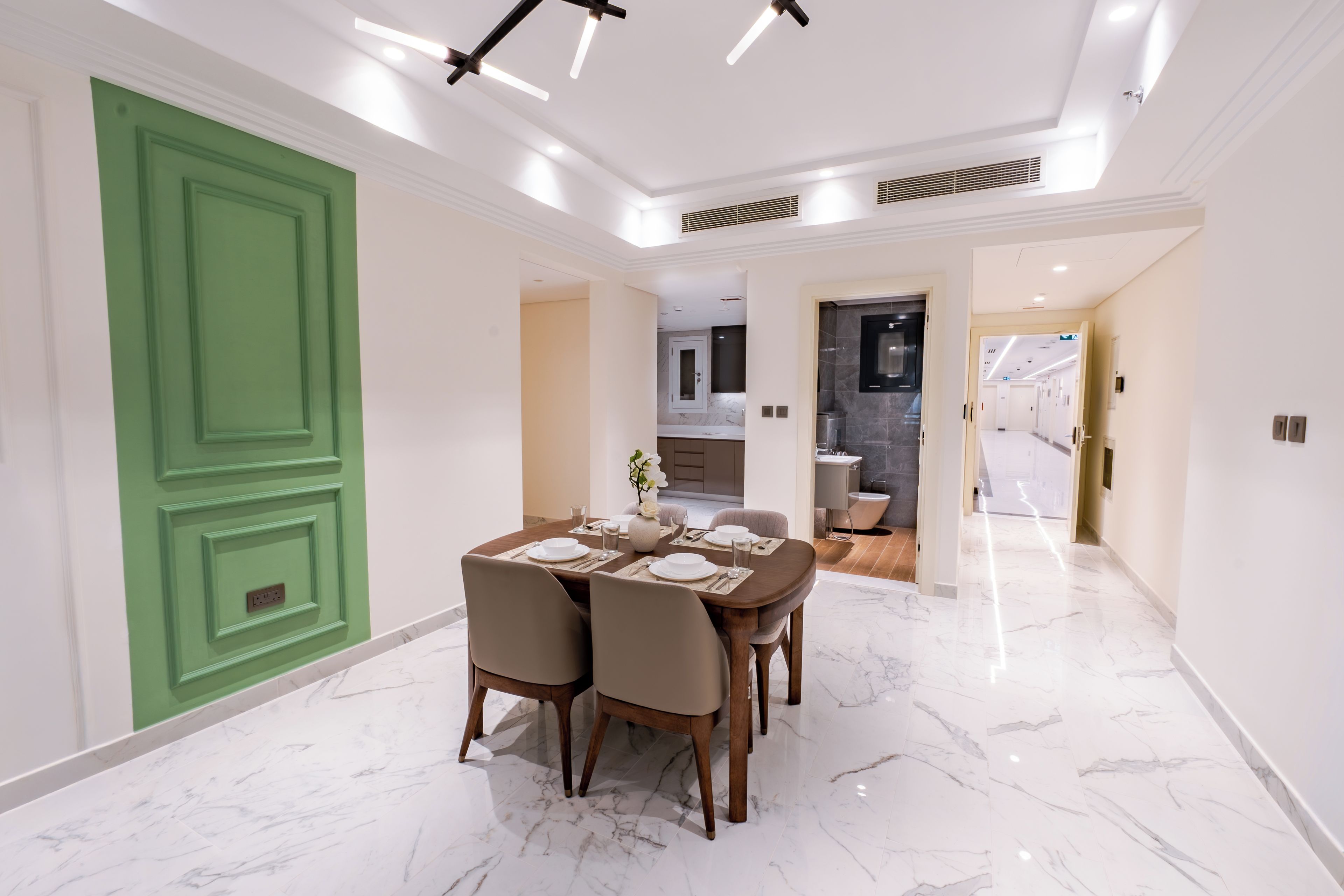 Uma sala de jantar com piso de mármore, uma porta verde e uma cozinha moderna ao fundo.