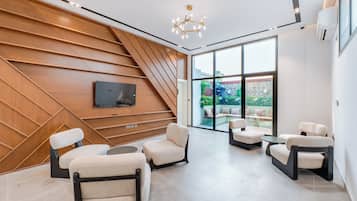 Um interior moderno com televisão fixada numa parede inclinada de madeira, área de estar com sofás e poltronas, e amplas janelas com vista para um jardim.