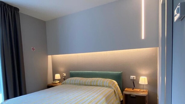 A hotel room with a bed, two bedside tables, a nightstand, a wall-mounted light, and a curtain. - Delta Park (Porto Viro)