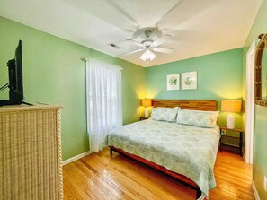 A bedroom with a bed, two bedside lamps, a ceiling fan, a television on a stand, and a wicker chair. - Emmas Beach Hut 2 KING BEDS 3 min to beach (Myrtle Beach)