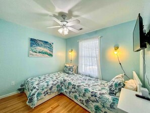 Un dormitorio con cama, ventilador de techo, un cuadro, ventana con cortinas y televisor montado en la pared.