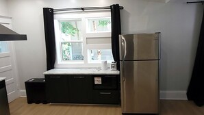 A compact kitchen with a stainless steel refrigerator, a sink, and a window with curtains. - Central Moncton 2BR | Walk, Pets & Parking (Moncton)