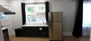 A compact kitchen with a stainless steel refrigerator, a sink, and a window with curtains. - Central Moncton 2BR | Walk, Pets & Parking (Moncton)