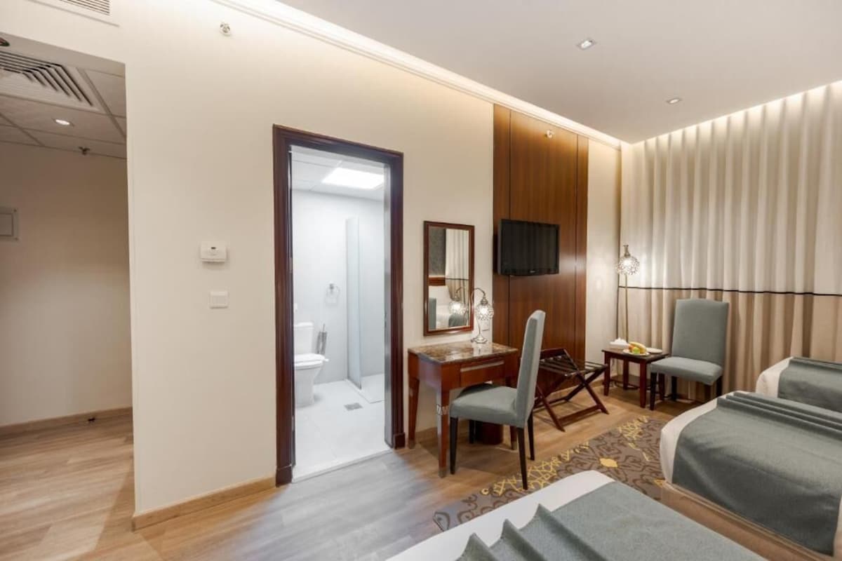 Quarto de hotel com uma cama, uma escrivaninha, uma cadeira, uma televisão e um banheiro visível por uma porta aberta.