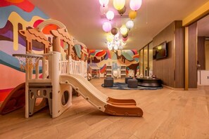 Un espace de jeux avec un toboggan et des structures d’escalade, un téléviseur fixé au mur et une fresque colorée sur le mur.